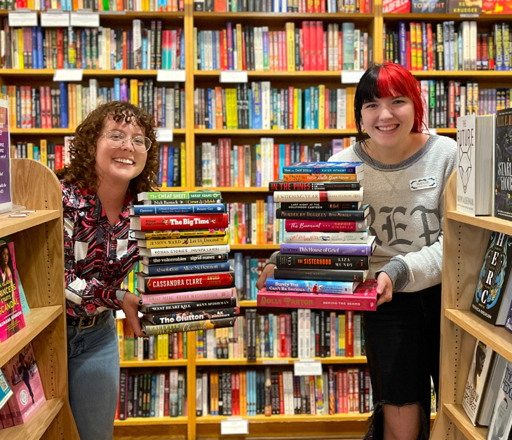 Booksellers Maddie and Lauren peek out from behind the stacks with piles of books in their hands